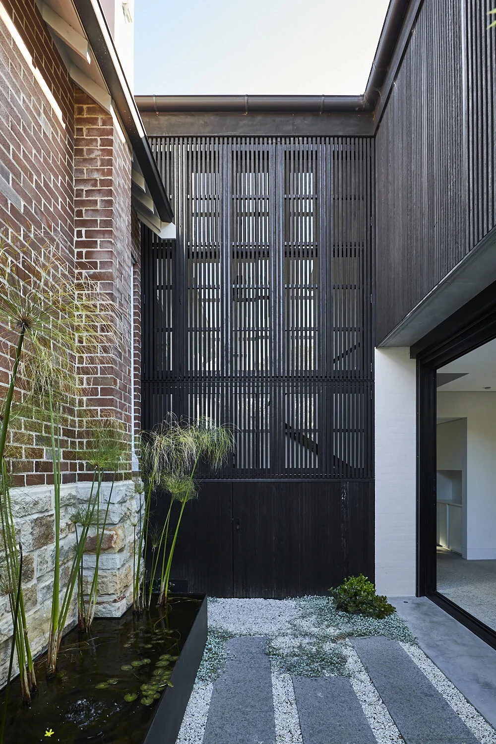 Modern outdoor courtyard with brick and black wooden walls, a water feature with plants, and a gravel pathway.