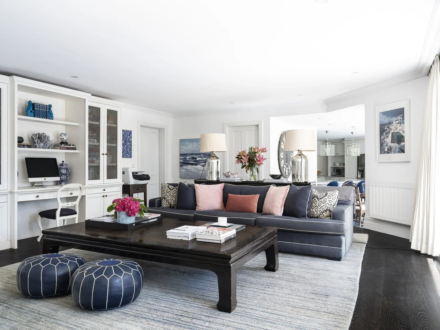 Bright open living room with dark hardwood floors, a light-colored area rug, a dark blue velvet sofa with pink and patterned throw pillows, a large wooden coffee table, decorative blue and pink ottomans, lamps, framed artwork, built-in white bookshel