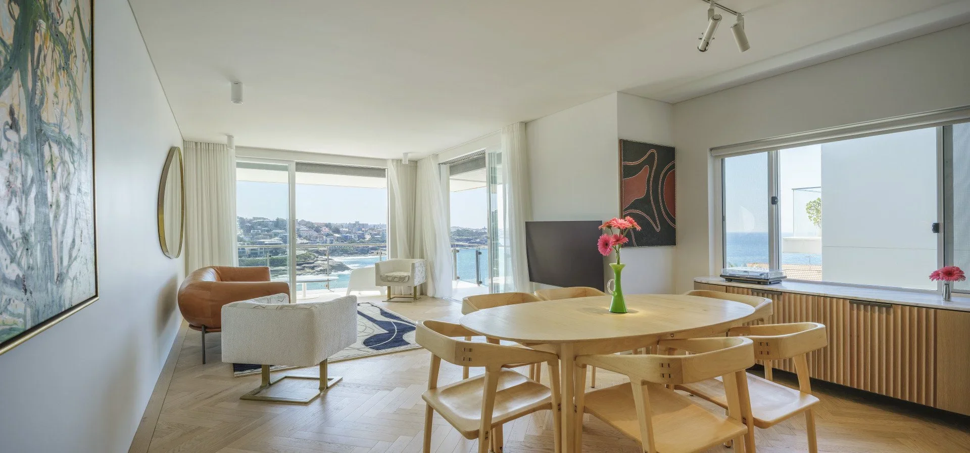 Living room with large windows showing a cityscape and ocean view, modern furniture including a round wooden dining table with pink flowers, a beige armchair, an orange sofa, a white armchair, a TV, and artistic wall decor.