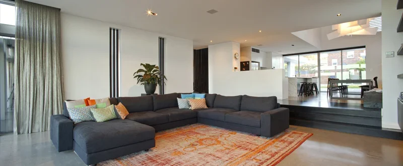 Modern living room with a large dark gray sectional sofa, colorful throw pillows, a potted plant, and an orange patterned rug, with an open view into the dining area and kitchen.