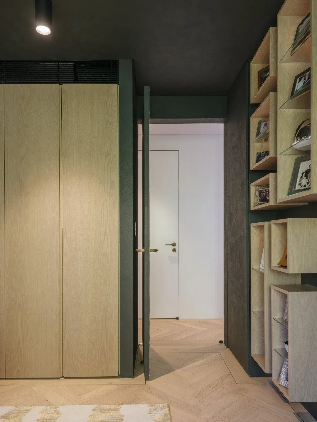 Interior view of a modern room with a wooden wardrobe on the left, a doorway with a black frame in the center, and a wall with wooden shelves holding photo frames and decorative items on the right.