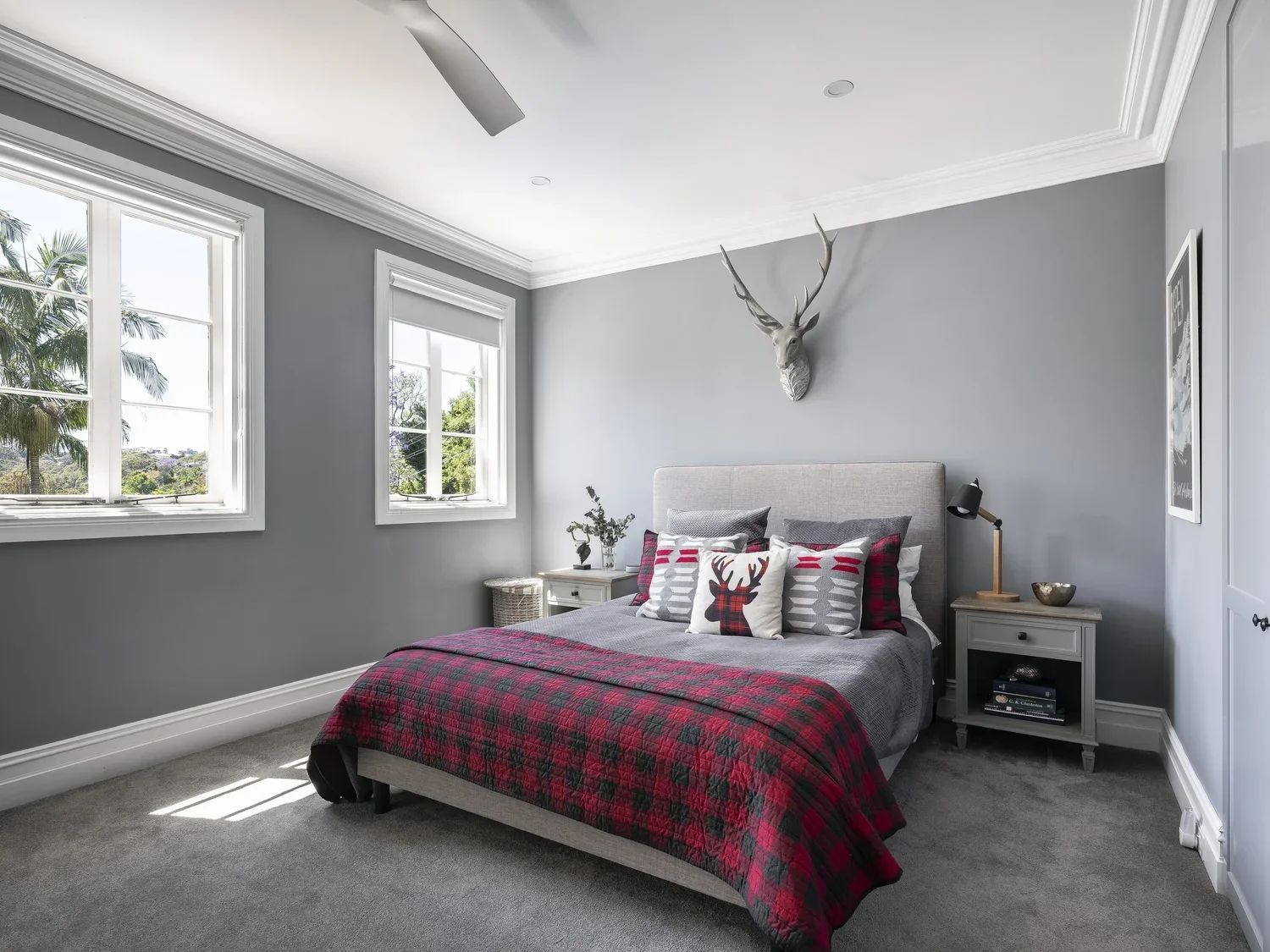 A bedroom with light gray walls, a gray upholstered bed with decorative pillows including one with a reindeer, a red plaid blanket, two nightstands with lamps, and a mounted deer head on the wall above the bed. There are three windows letting in natu