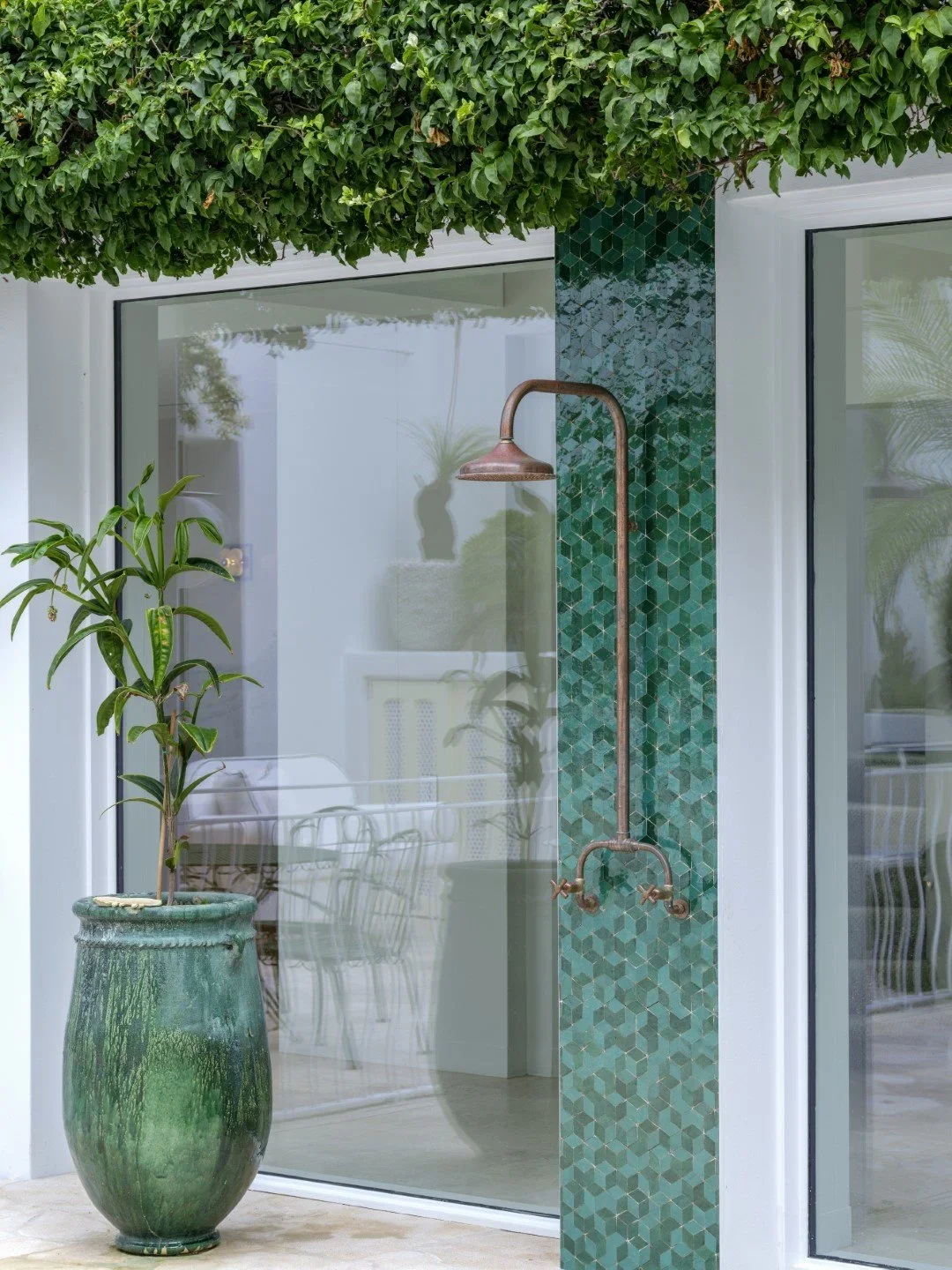 Outdoor shower with green mosaic tile accent wall, copper showerhead, and large potted plant, with sliding glass door and greenery overhead.
