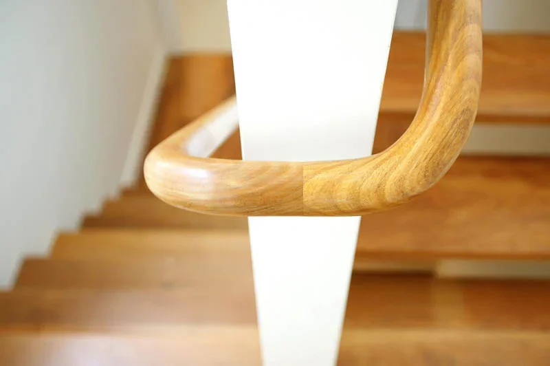 Close-up view of a wooden stair handrail attached to a white painted support post, with stairs visible in the background.