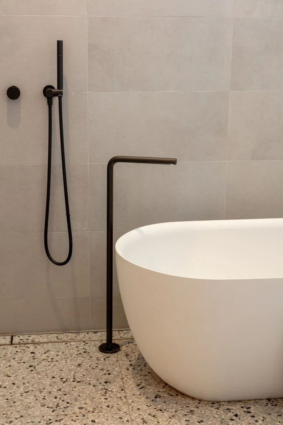 Modern bathroom with a white freestanding bathtub, a matte black standing faucet, and a black handheld shower with a hose mounted on large light gray wall tiles, and a speckled terrazzo floor.