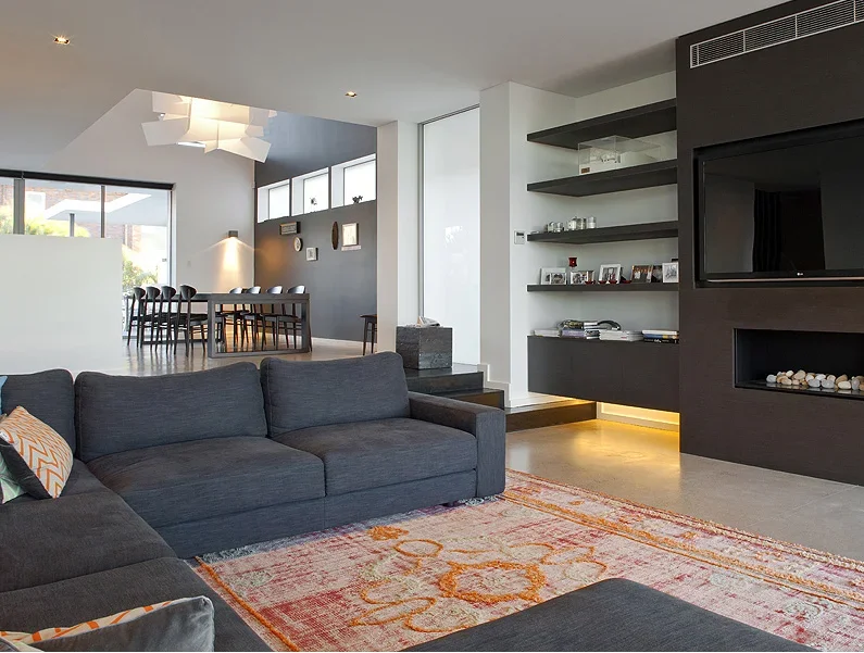 Modern living room with dark gray sofa, colorful rug, and black fireplace built into dark wall, open shelves with framed photos, and a dining area with black chairs and a large window.