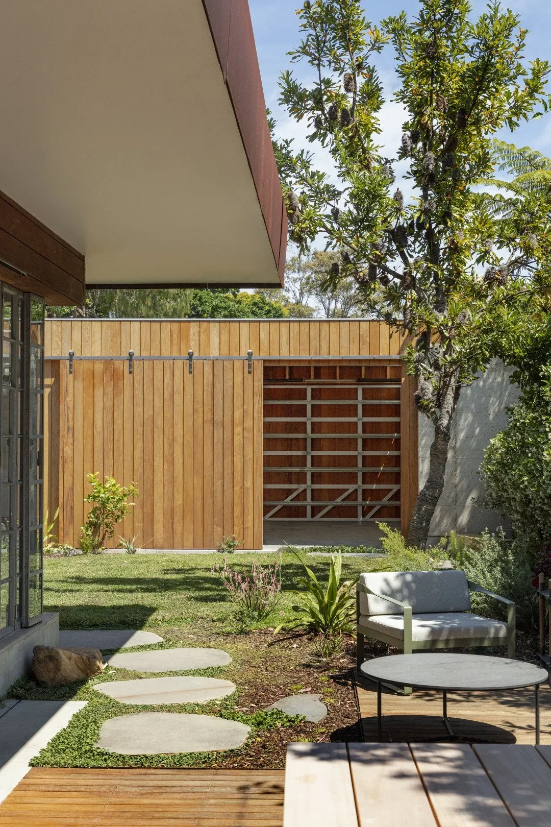 A modern backyard patio with a wooden privacy fence, a green lawn with stepping stones, a white cushioned outdoor sofa near a round black coffee table, and trees and plants providing shade and greenery.