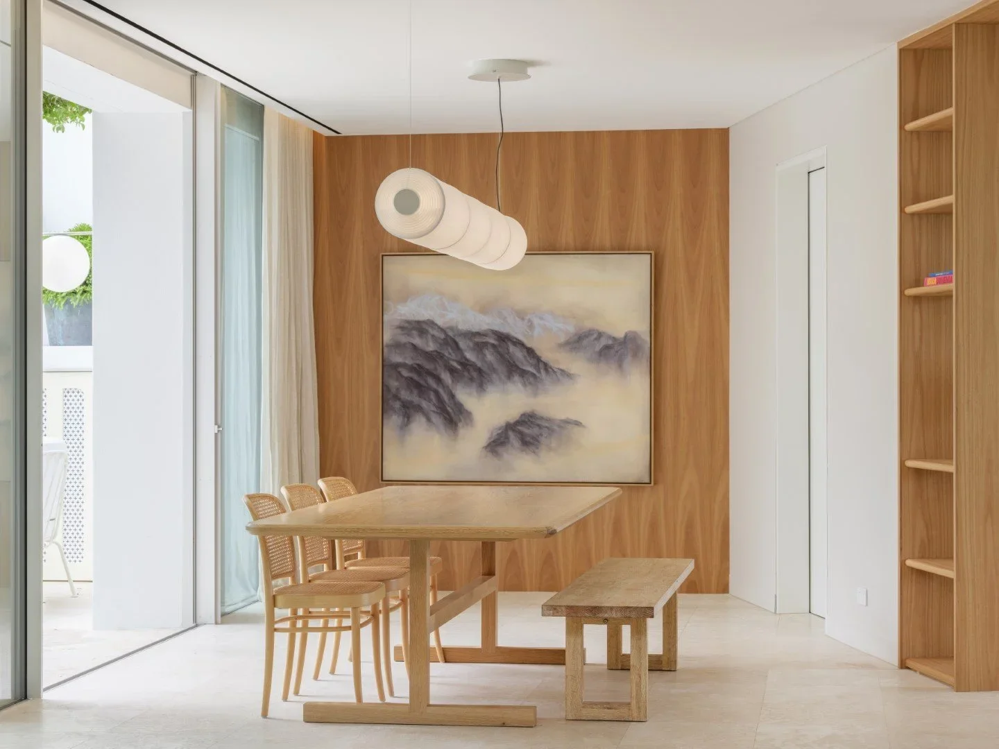 Modern dining area with wooden table, matching chairs and a bench, large abstract mountain painting, wooden wall panel, pendant light, and a sliding glass door with a view of a balcony.