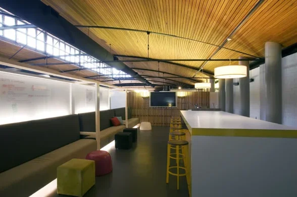 Modern lounge area with long sofa, colorful ottomans, yellow barstools, and wooden ceiling in a contemporary space.