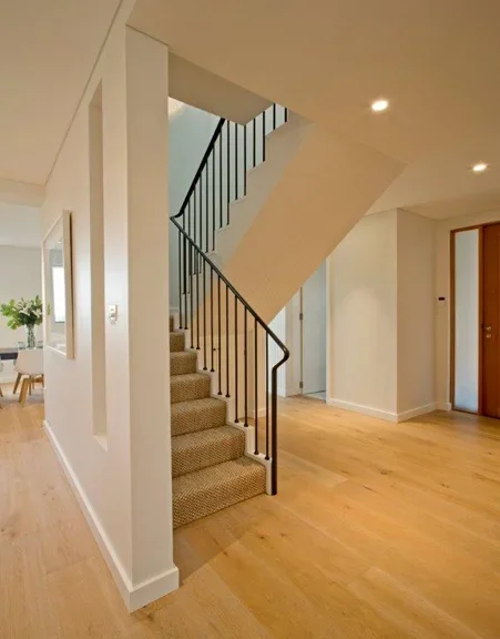 Empty living space with wooden floors, a staircase with carpeted steps, black railing, and white walls.