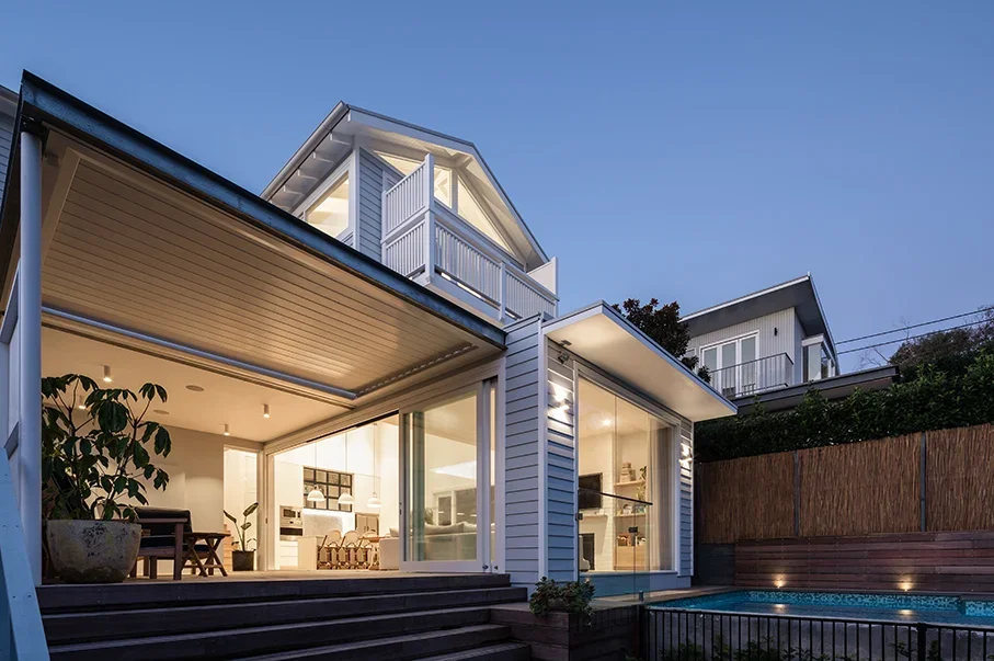 Modern multi-story house with large glass windows, a wooden deck, a small swimming pool, and outdoor lighting at dusk.