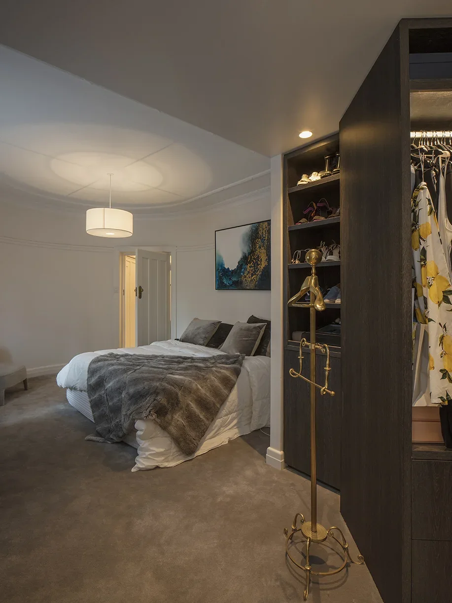 A bedroom with a bed that has gray pillows and a fur blanket, a wall painting, a clothing closet with shoes, and a standing coat rack, all in a modern interior with beige carpet and white walls.