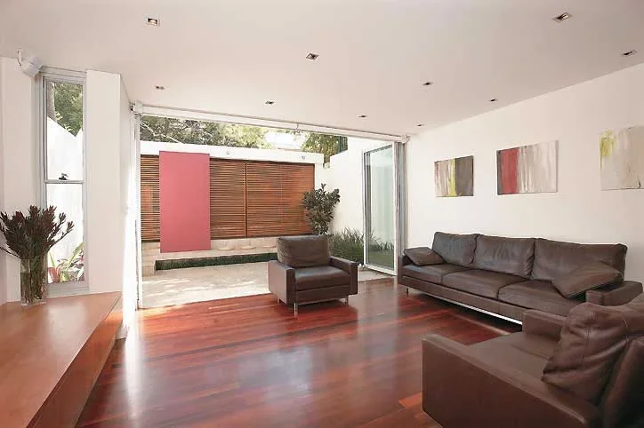 Modern living room with dark gray sofas, wooden flooring, and large glass sliding doors leading to an outdoor patio with plants and a wooden privacy screen.