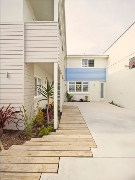 Modern two-story house with white and blue exterior, small garden with plants, wooden pathway, and concrete driveway in a small courtyard.