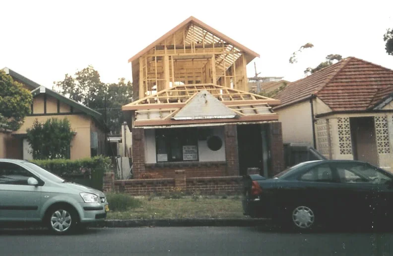 A house under construction with a wooden framework and a pitched roof, parked cars on the street in front, and neighboring houses on either side.