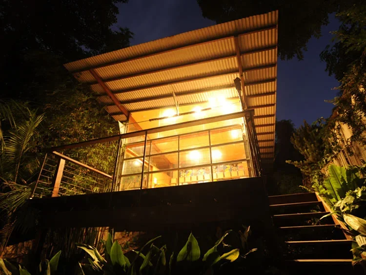 A house with large glass windows and a metal roof in a lush green setting at night, lit from inside.