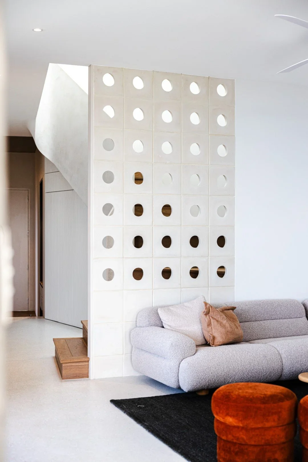 Living room with a modern beige sofa, two brown pillows, black rug, orange pouf, white wall with a decorative partition with circular cut-outs, and ceiling fan.