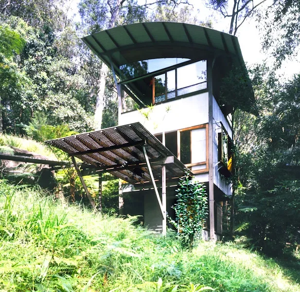 Unique multi-level house with a green curved roof, elevated on stilts, surrounded by lush greenery in a forest setting.