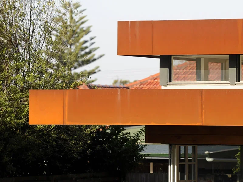 A modern building with orange exterior walls and a large window on the upper floor, with trees and a rooftop visible in the background.