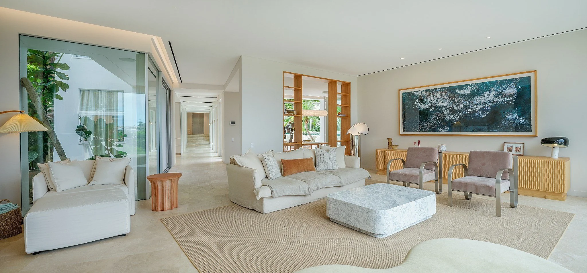 Living room with cream-colored sofa, marble coffee table, white armchair, fireplace, sliding glass door showing greenery, and minimalistic decor.