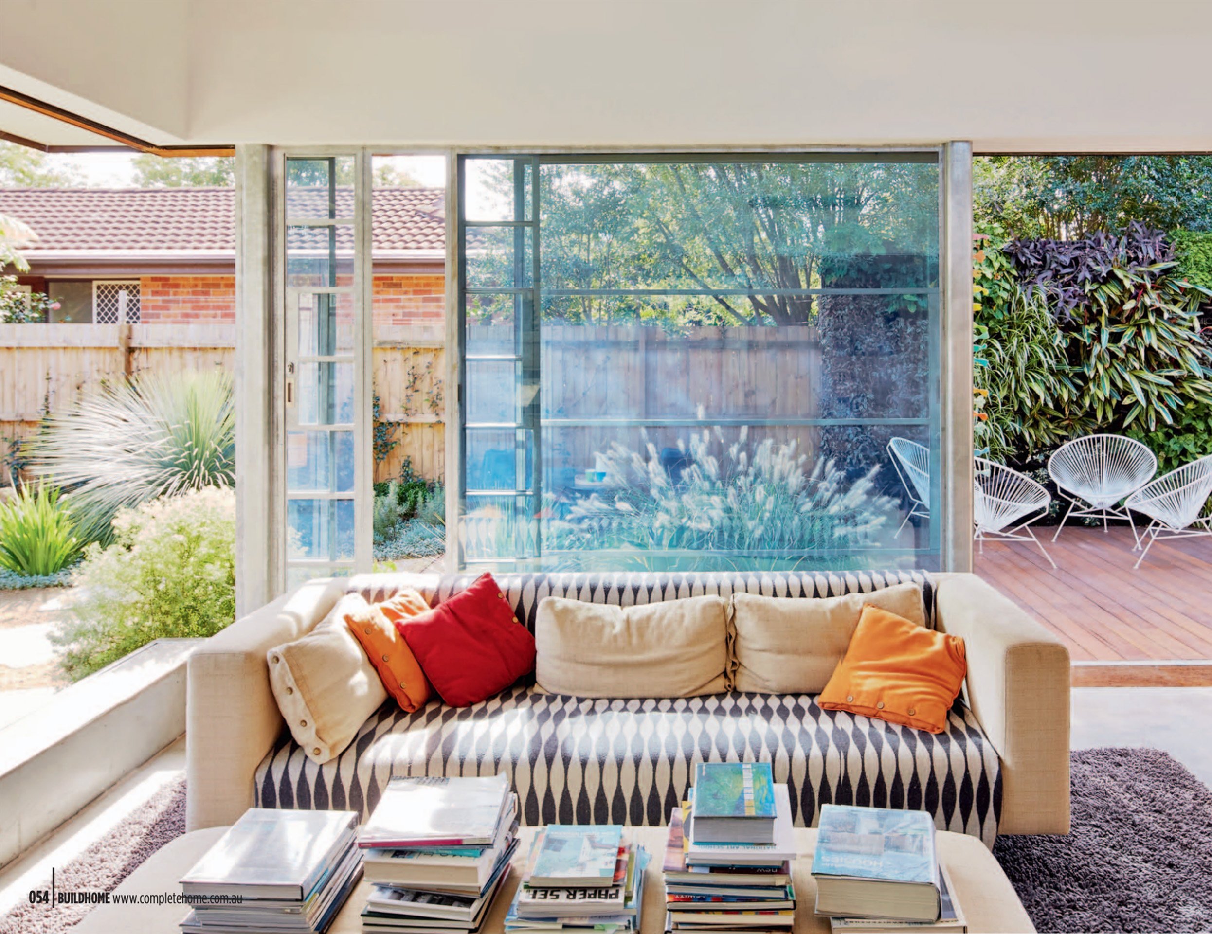 Living room with large windows looking out onto a backyard featuring outdoor chairs and lush greenery.
