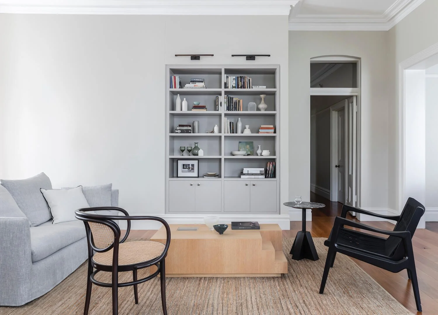 Modern living room with a light gray sofa, black and wicker chairs, a wooden coffee table, a small side table with a glass, a bookshelf with decorative items, and hardwood flooring.