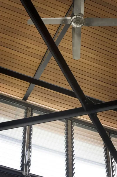 Ceiling fan with wooden blades and metal frame, geometric black metal beams crossing, and large windows with horizontal blinds.
