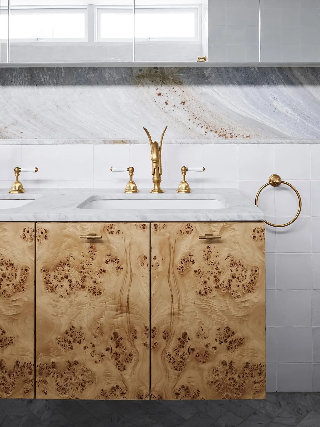 Close-up of a bathroom vanity with a white marble countertop, three gold faucet handles, and a gold faucet. The vanity has light-colored wood cabinets with a burl wood pattern. Behind the sink is a white tiled wall, and a gold towel ring is mounted o
