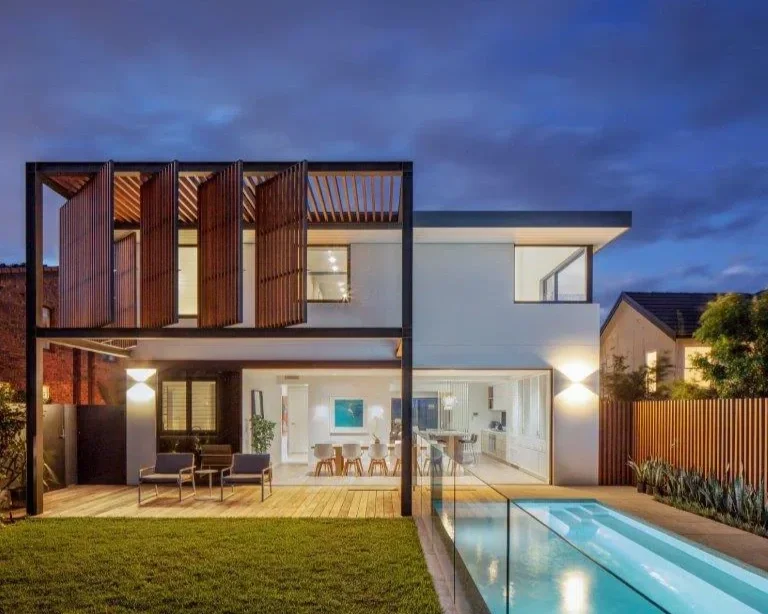 Modern two-story house with a large glass sliding door, a backyard with a swimming pool, outdoor seating area, and a wooden privacy fence, illuminated at dusk.