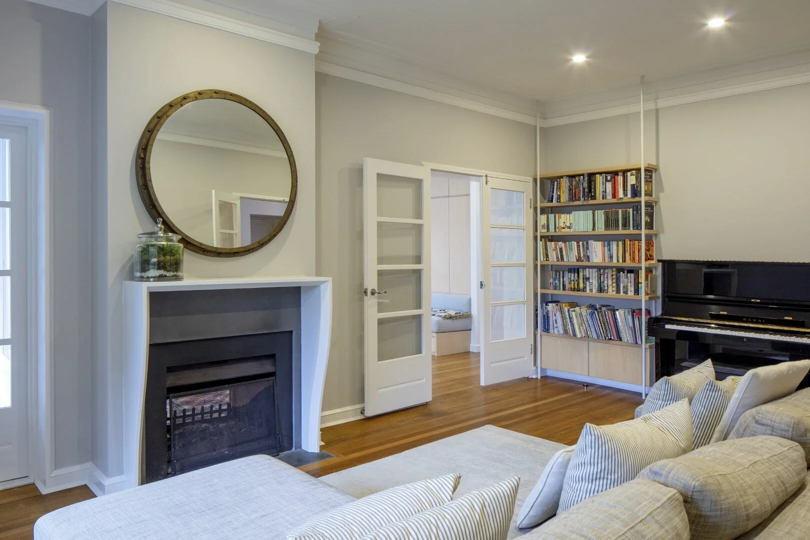 Living room with beige sofa, striped pillows, white fireplace with mirror above, wooden bookshelf filled with books, upright black piano, wooden floors, glass-paneled double doors leading to another room, and a small window.