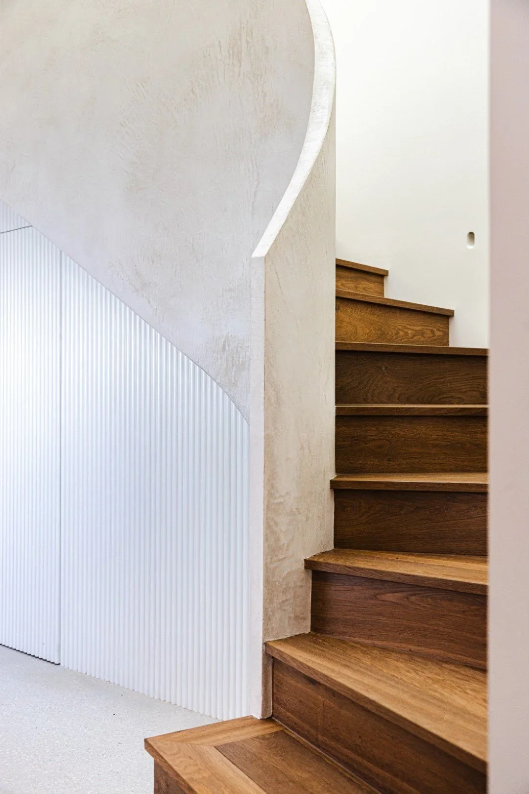 Close-up of a wooden staircase with beige textured wall panels and a curved white wall.