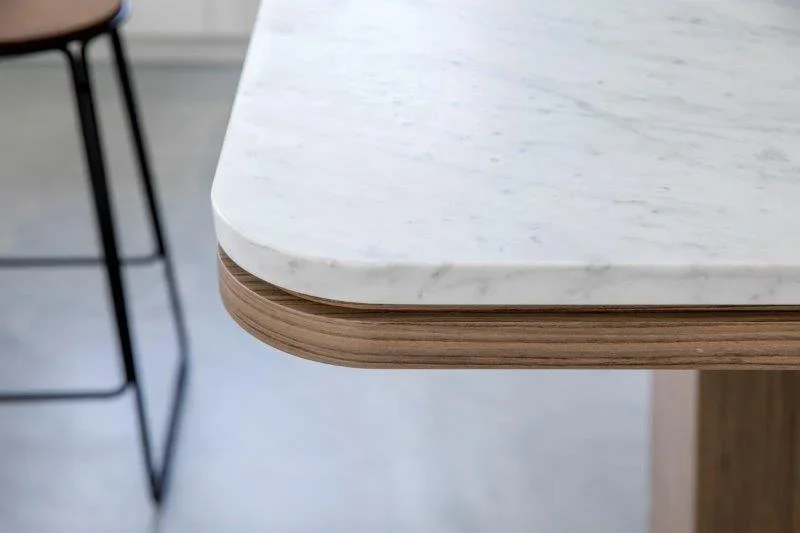 Close-up of a corner of a table with a white marble top and wooden edging, with a black metal chair leg partially visible in the background.