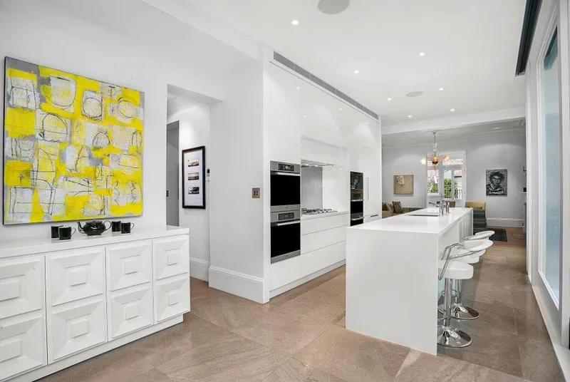 Modern, white kitchen with built-in ovens, a large island with bar stools, and artwork on the walls.