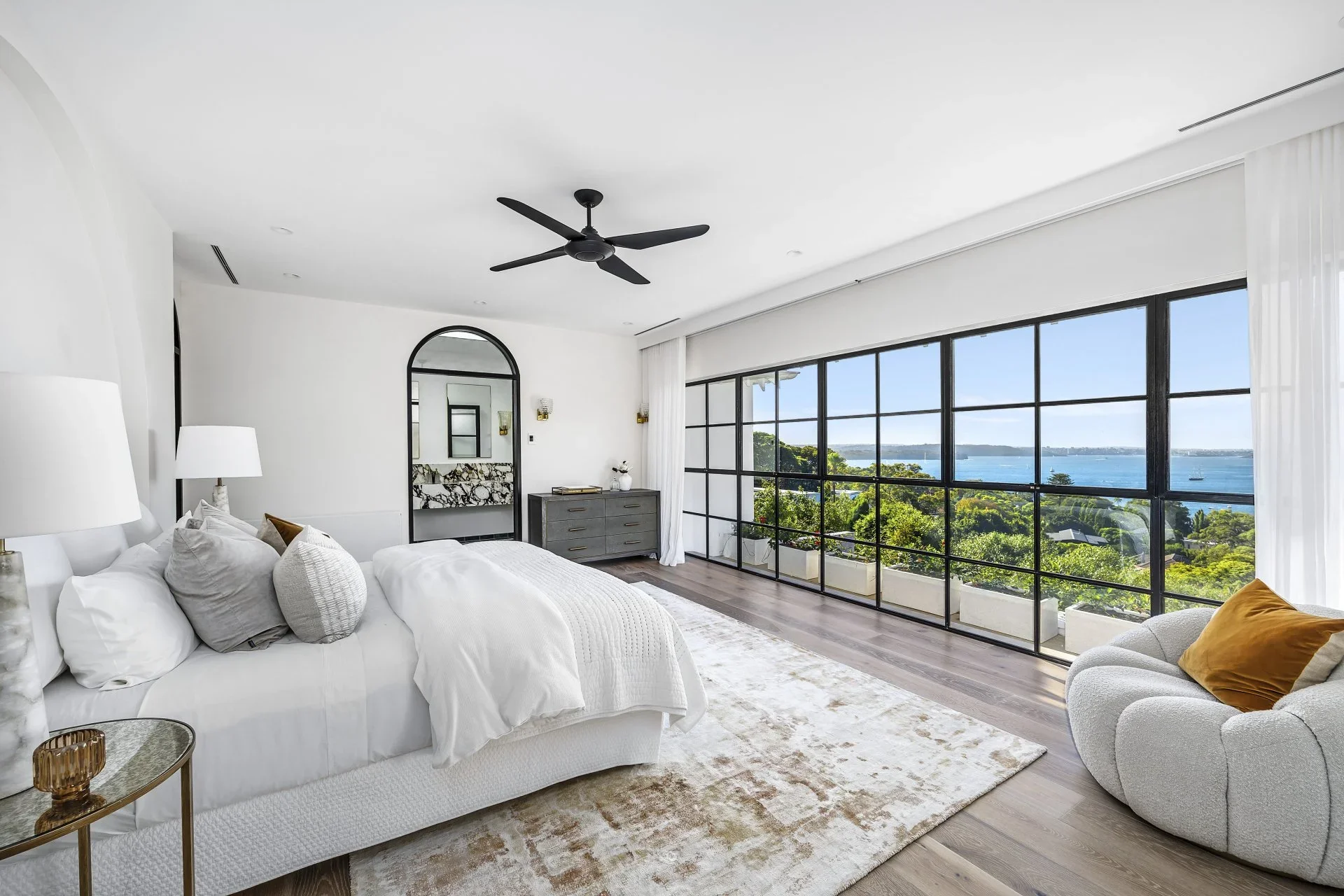 Modern bedroom with large window view of water and greenery, white bed, gray dresser, armchair with yellow pillow, ceiling fan, white walls, hardwood floor, and minimal decor.