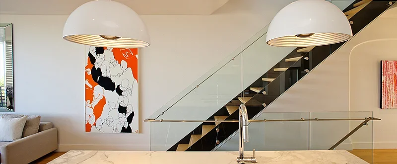 Modern living room with a staircase, glass railing, and contemporary artwork, with a white pendant light hanging above.
