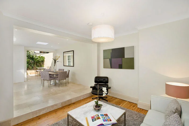 Living room with white walls, hardwood floor, a black lounge chair, a white sofa, a marble coffee table with an open book and plant, and a framed artwork on the wall. Adjacent dining area with a table and chairs, visible through an open space, with o
