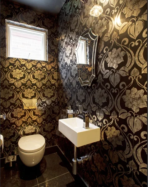 Small bathroom with black and gold floral wallpaper, a small white sink, a toilet, a mirror, and two small windows.