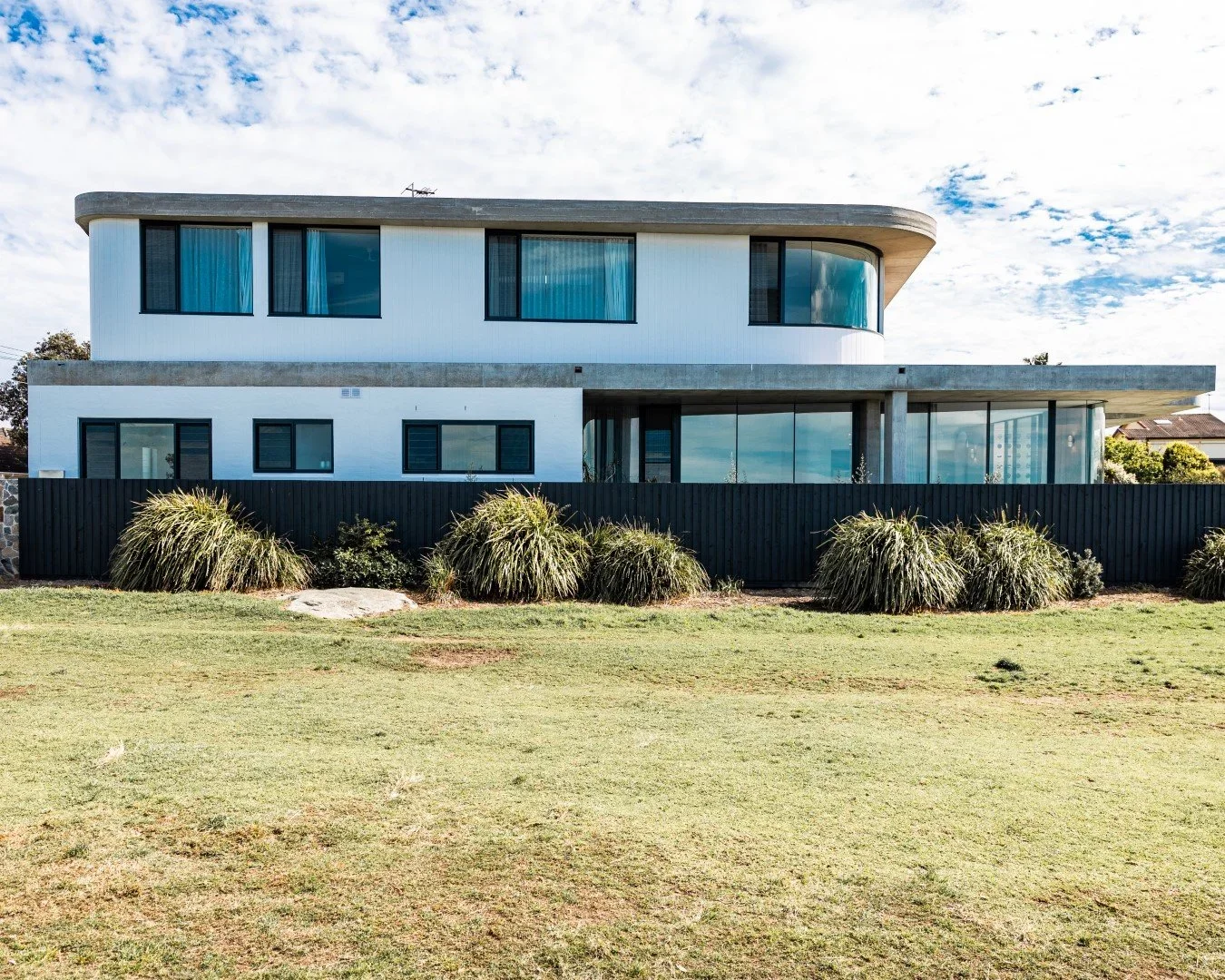 Modern two-story house with large glass windows and a white exterior, surrounded by a black wooden fence and landscaped yard with bushes and grass.