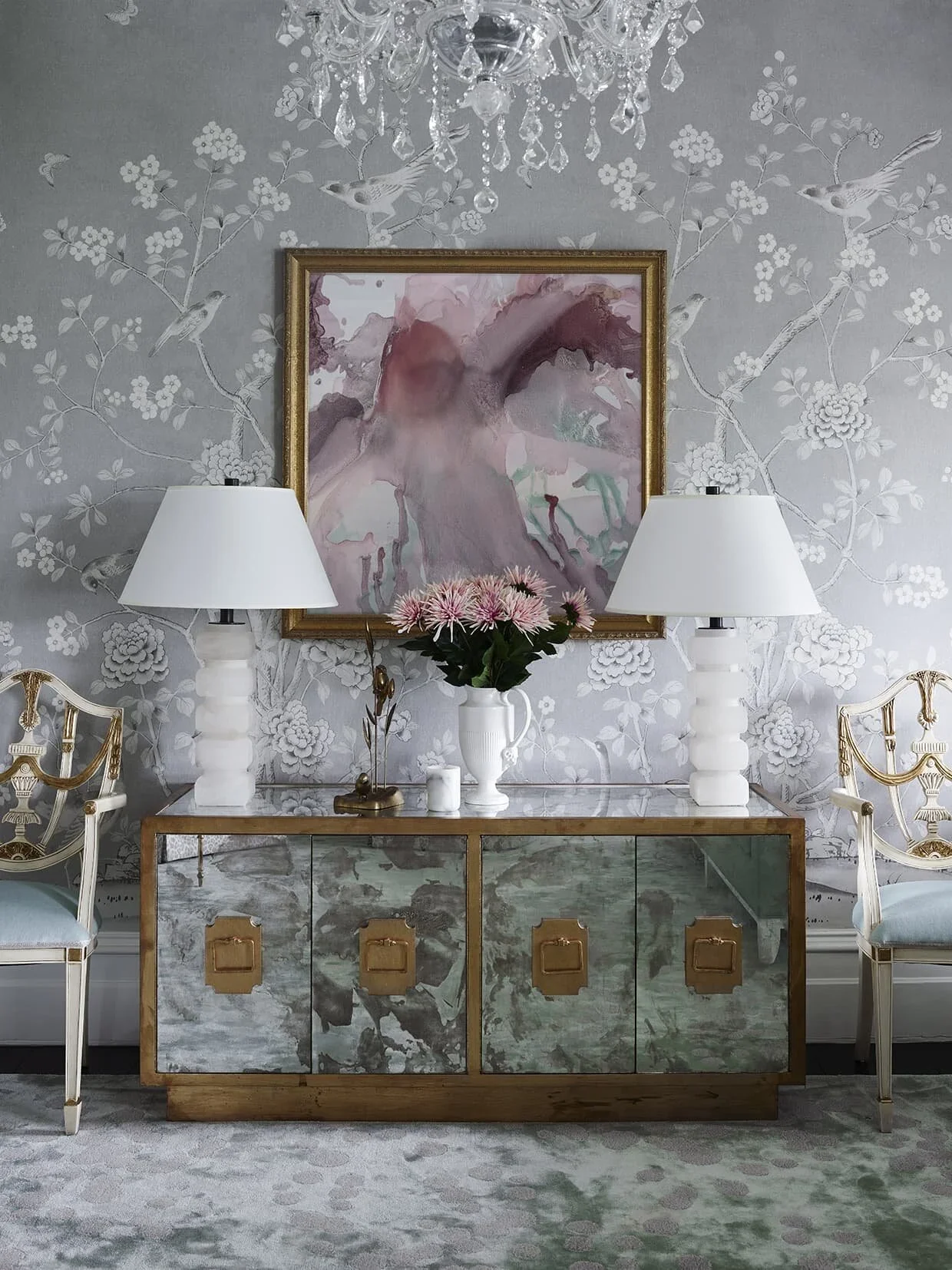 A decorated living room with a grey floral wallpaper, a chandelier, a gold-framed painting, a mirrored sideboard with a vase of pink flowers, two white table lamps, and two elegant chairs with gold accents.