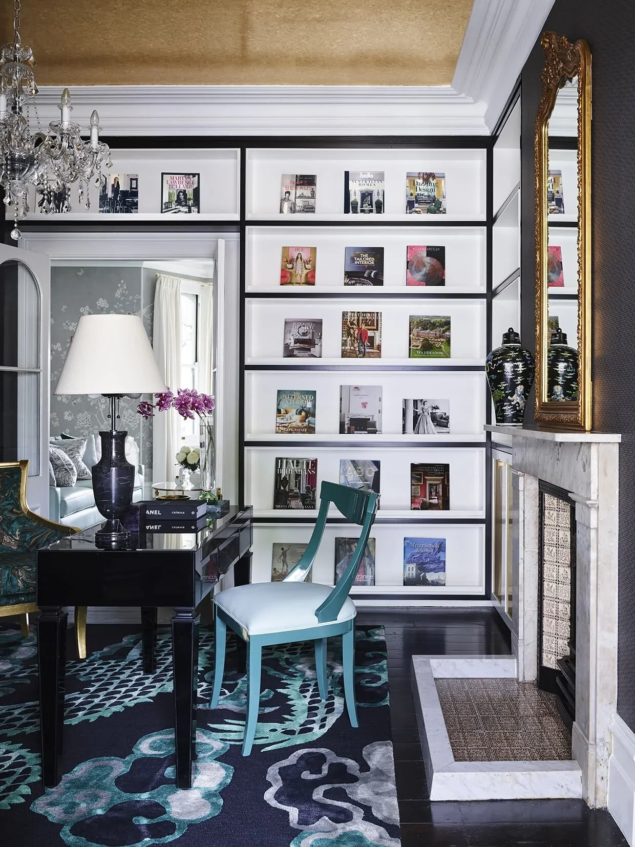 Interior of a living room with a bookshelf, a fireplace, a mirror, a black table with a lamp, a teal chair, a patterned rug, and decorative vases.