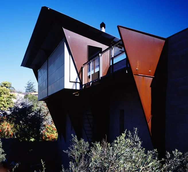 Modern house with angular design features rust-colored and black panels, with large windows and a balcony, set against a clear blue sky with surrounding greenery.