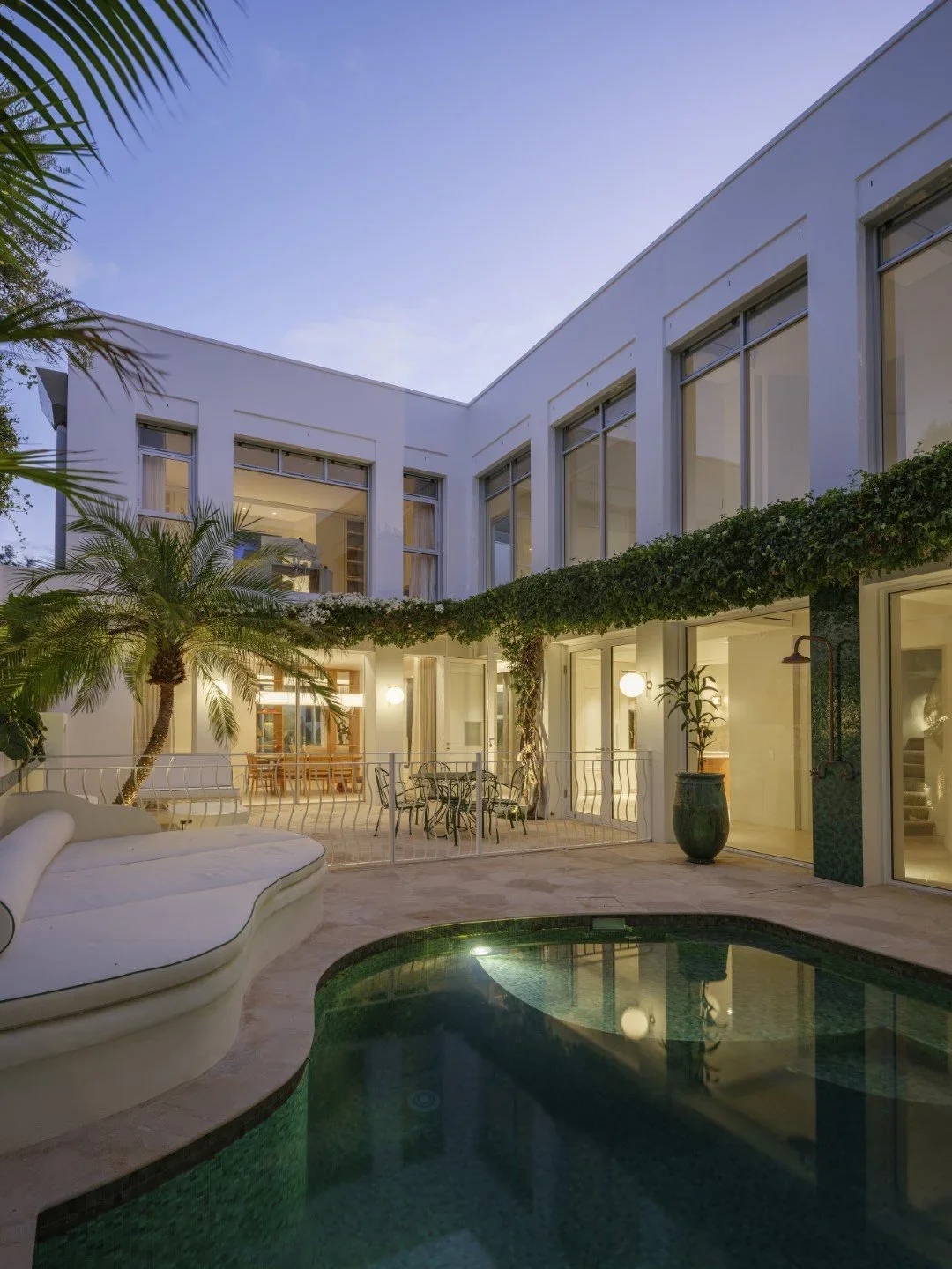 A modern white residential building with large windows, outdoor seating area, a swimming pool, lush green plants, and string lights, during dusk.