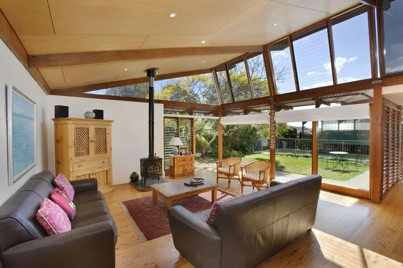 Living room with leather sofas, wooden cabinet, wood stove, coffee table, two chairs, and large windows showing a backyard with green grass and trees.