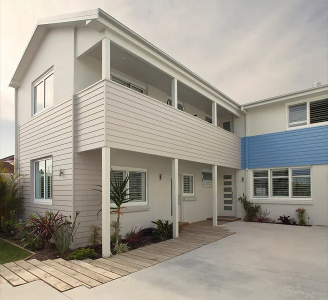 Exterior of a modern, multi-story house with white siding, a blue accent on the second floor, and large windows. There is a small landscaped area with plants and a wooden walkway leading to the entrance.