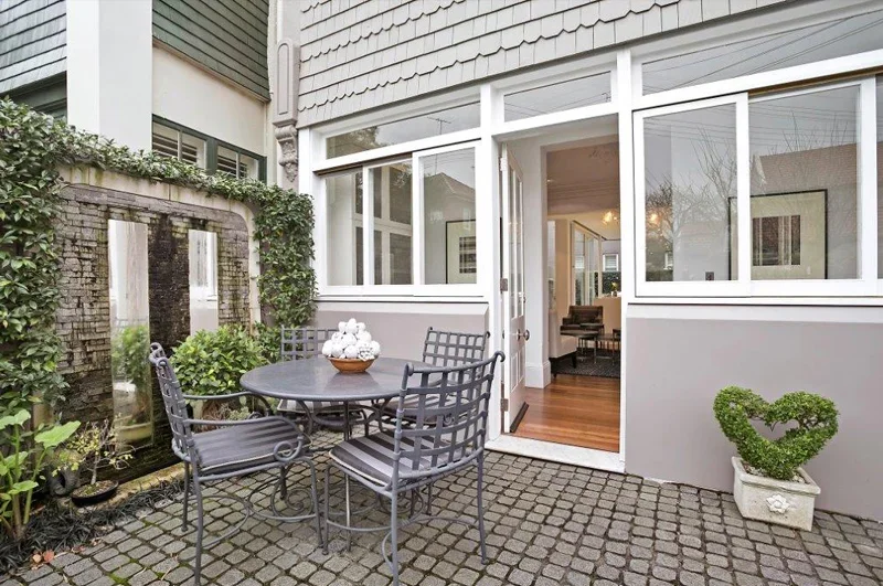 Outdoor patio with a round metal table and four matching chairs, decorated with a bowl of white flowers. The patio has a cobblestone floor and is adjacent to a house with large windows and a sliding glass door leading inside. There is greenery along 
