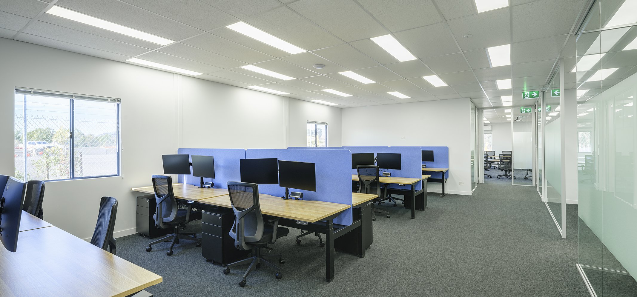 Empty modern office space with desks, chairs, and computer monitors. Bright lighting, large windows, and glass partitions. Signage indicating emergency exits.