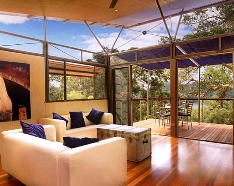 A cozy living room with a white sofa and blue pillows, opening to a deck with outdoor seating and a view of trees and a blue sky.