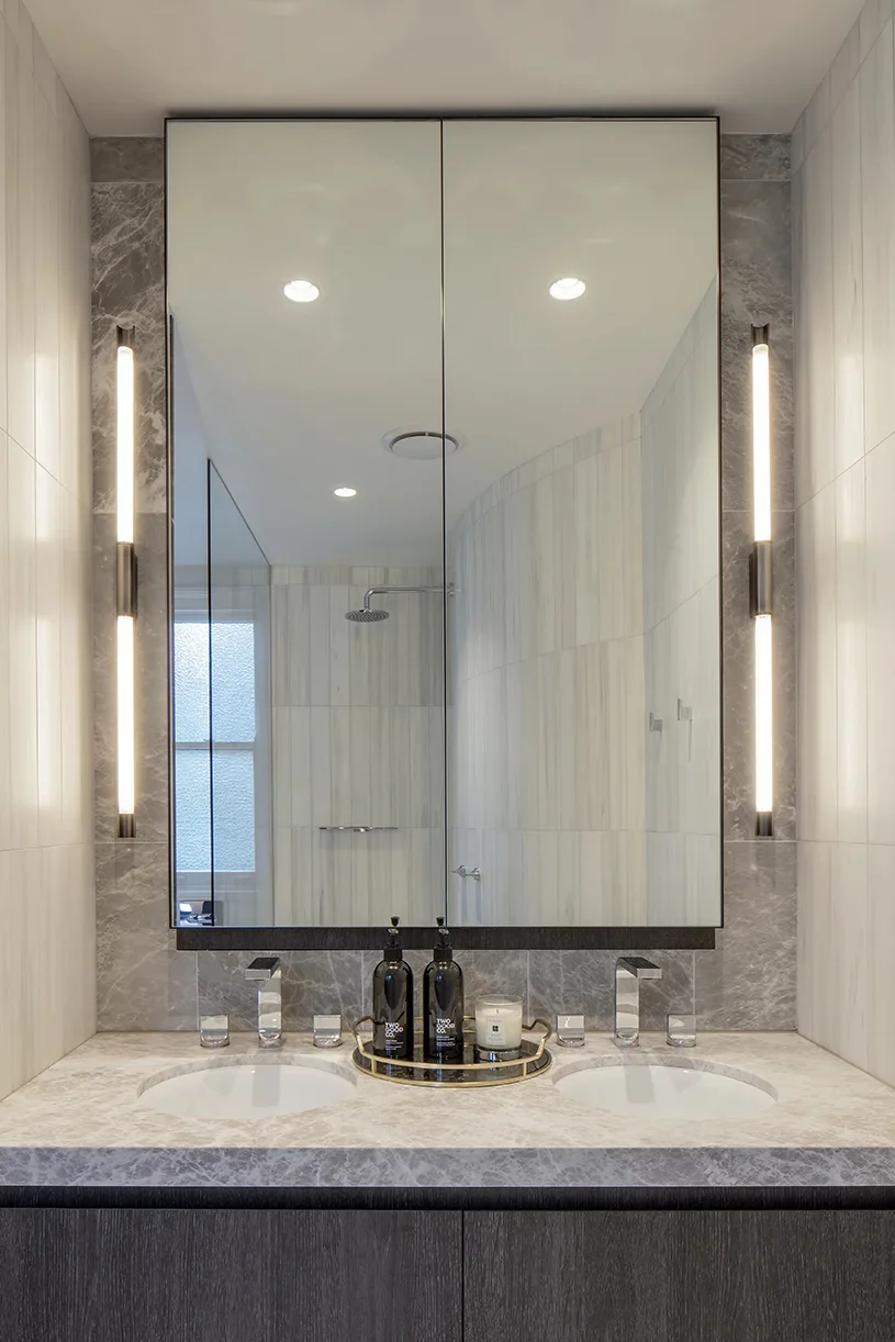 Modern bathroom vanity with a double sink, large mirror, and vertical light fixtures on either side, featuring a marble countertop and a shower visible through the mirror reflection.