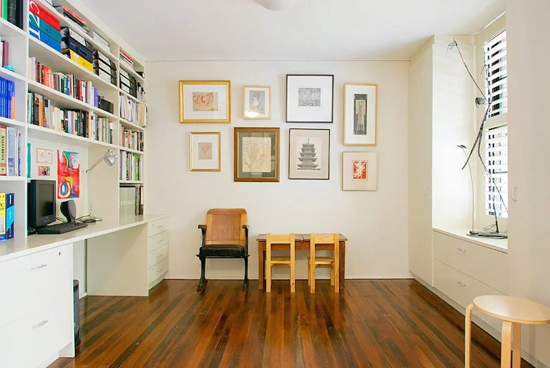 Living room with white wall decorated with framed art, a large white bookshelf filled with books and boxes, a small desk with a computer and speaker, a wooden chair, two small wooden chairs, and a window with shutters and a tall plant.