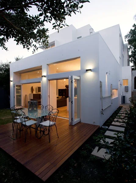 Modern white house with large glass sliding doors opening to a wooden deck with a small dining table and metal chairs, surrounded by greenery.